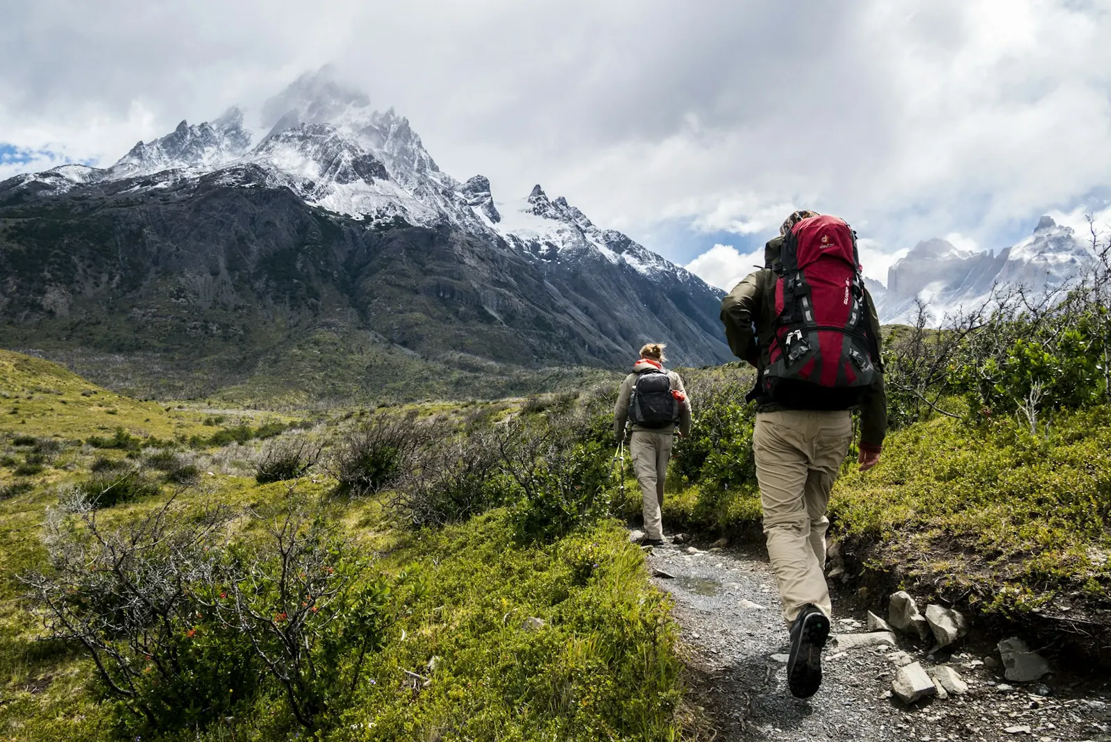 Two hikers walking along a trail towards snowy mountains