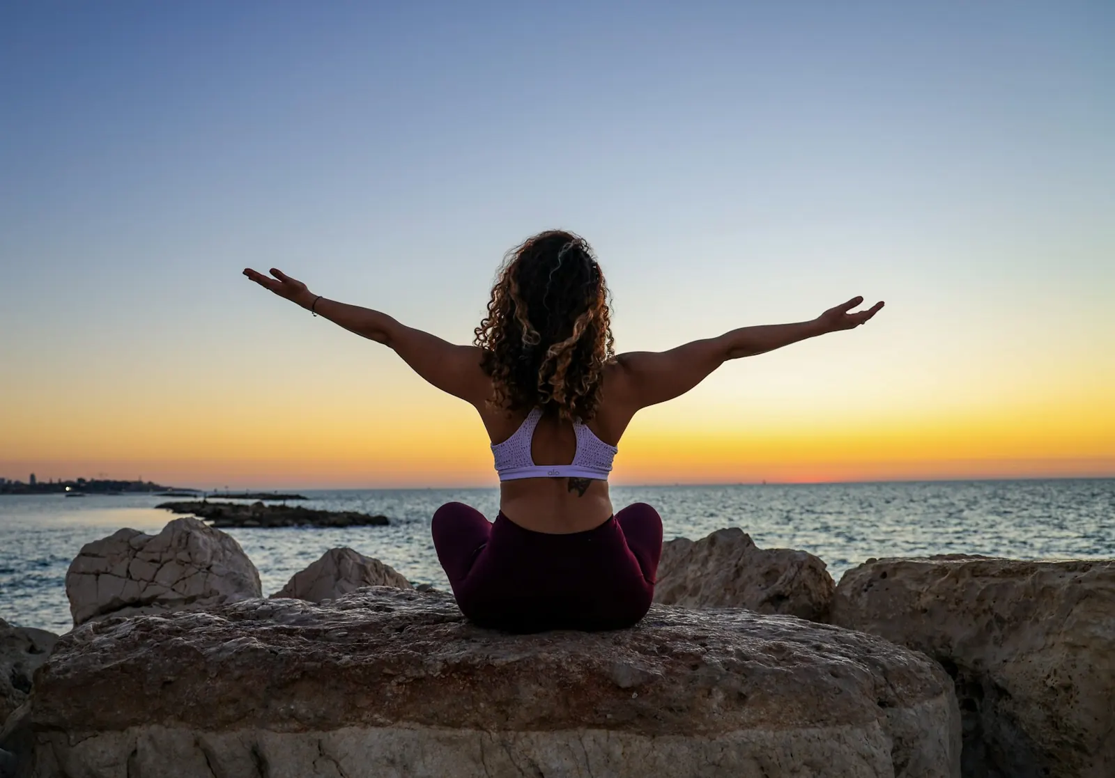 Woman sitting on rocks by the sea at sunset with arms stretched out