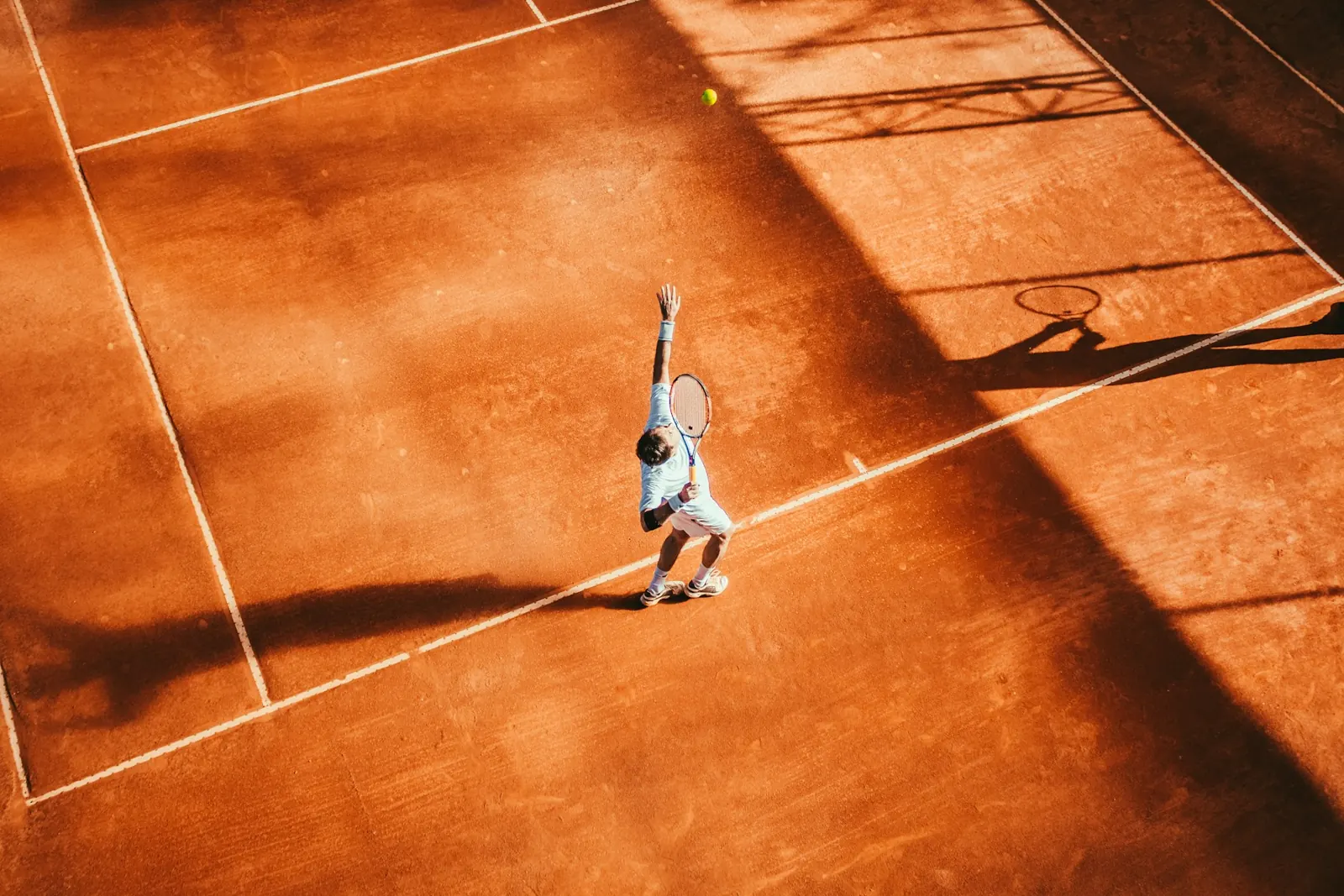 Tennis player tossing the ball to serve on an orange clay court, seen from above