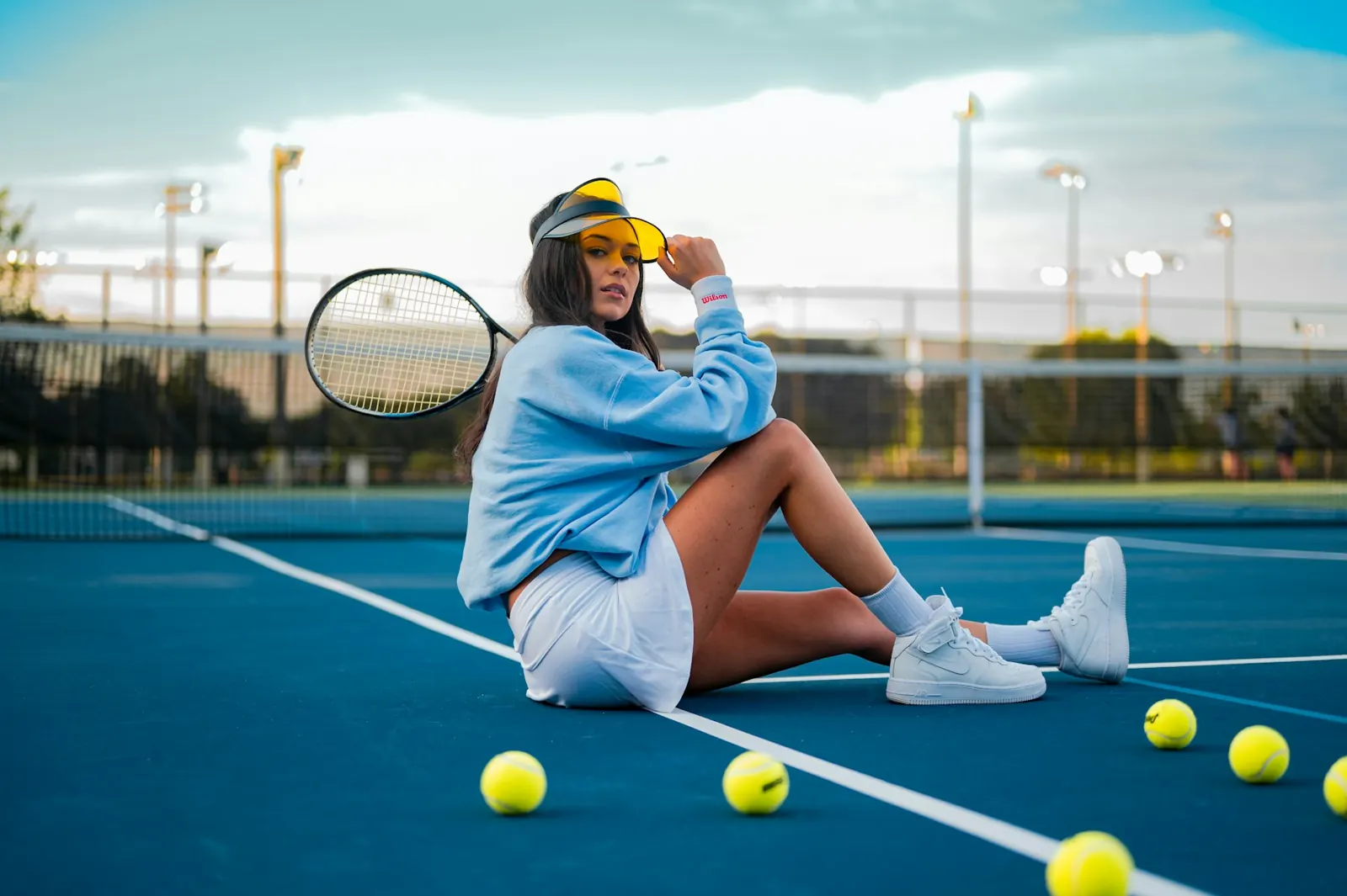 Tennis player sitting on a blue court surrounded by tennis balls, looking thoughtful